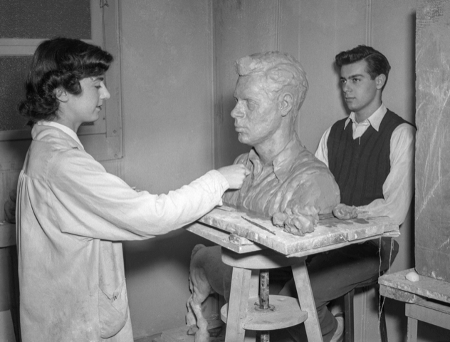 Kaish photo black and white photo of a woman on the left with a white coat on with dark hair, sculpting a bust with a model posing at far right
