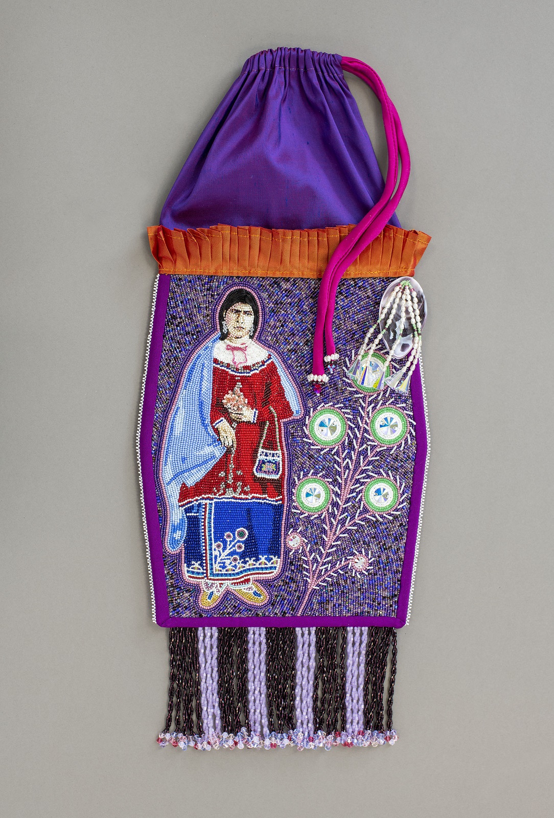 Purple drawstring pouch with tassels at bottom, fringe at the upper half, and beaded woman wiht a red shirt and blue skirt next to beaded floral