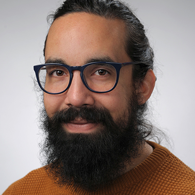 Victor Rivera Portrait portrait of a male with dark hair pulled back and a beard. wearing black glasses and wearing a brown sweater