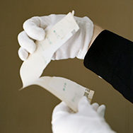 Color photo of a person's arm in a black shirt, and hand in a white glove holding open a sheet of paper being ripped in half