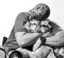 Black and white image of a man sitting in a wheelchair, hugging a service dog