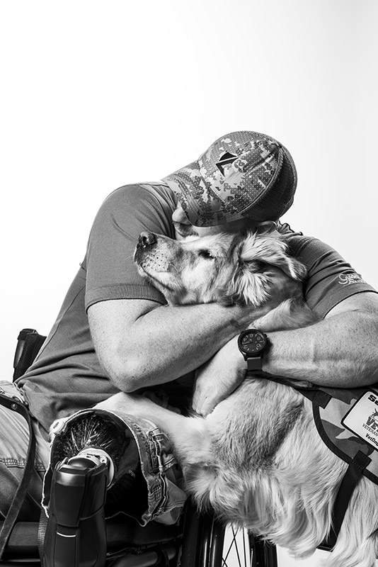Black and white image of a man sitting in a wheelchair, hugging a service dog