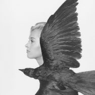 Black and white portrait of a female, in profile, with a crow in front of her, wings spread out.