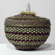 Collaborative hand-woven round, squat shaped basket bottom (b) and top (a), light green with thin bands of purple; the basket top has a central medallion shape consisting of bright light green-dyed moose hair and a pale bone-colored hand-carved moose antler sculpture of a robed figure that stands about 2" tall (carved by Stonehorse Goeman).