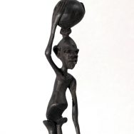 Black carved figure with a water jug on top of head, arm holding it steady.