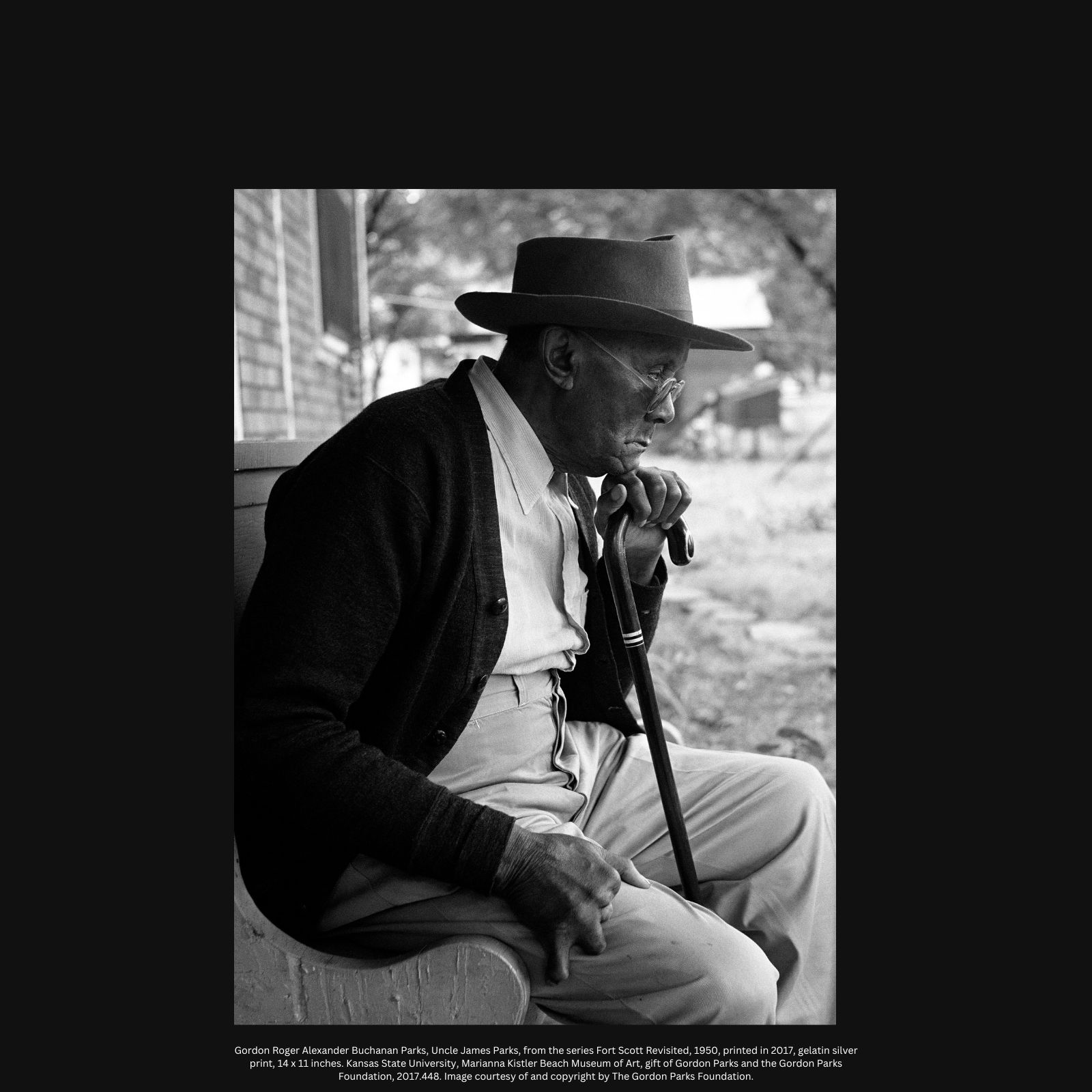 Black and white photo of an elderly male from the side wearing a hat, sitting down, holding on to a cane