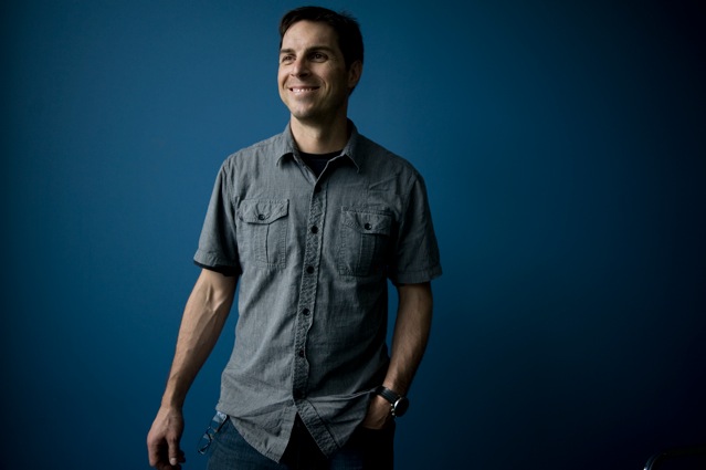 Portrait of a male, standing wearing a short sleeve gray shirt, blue background